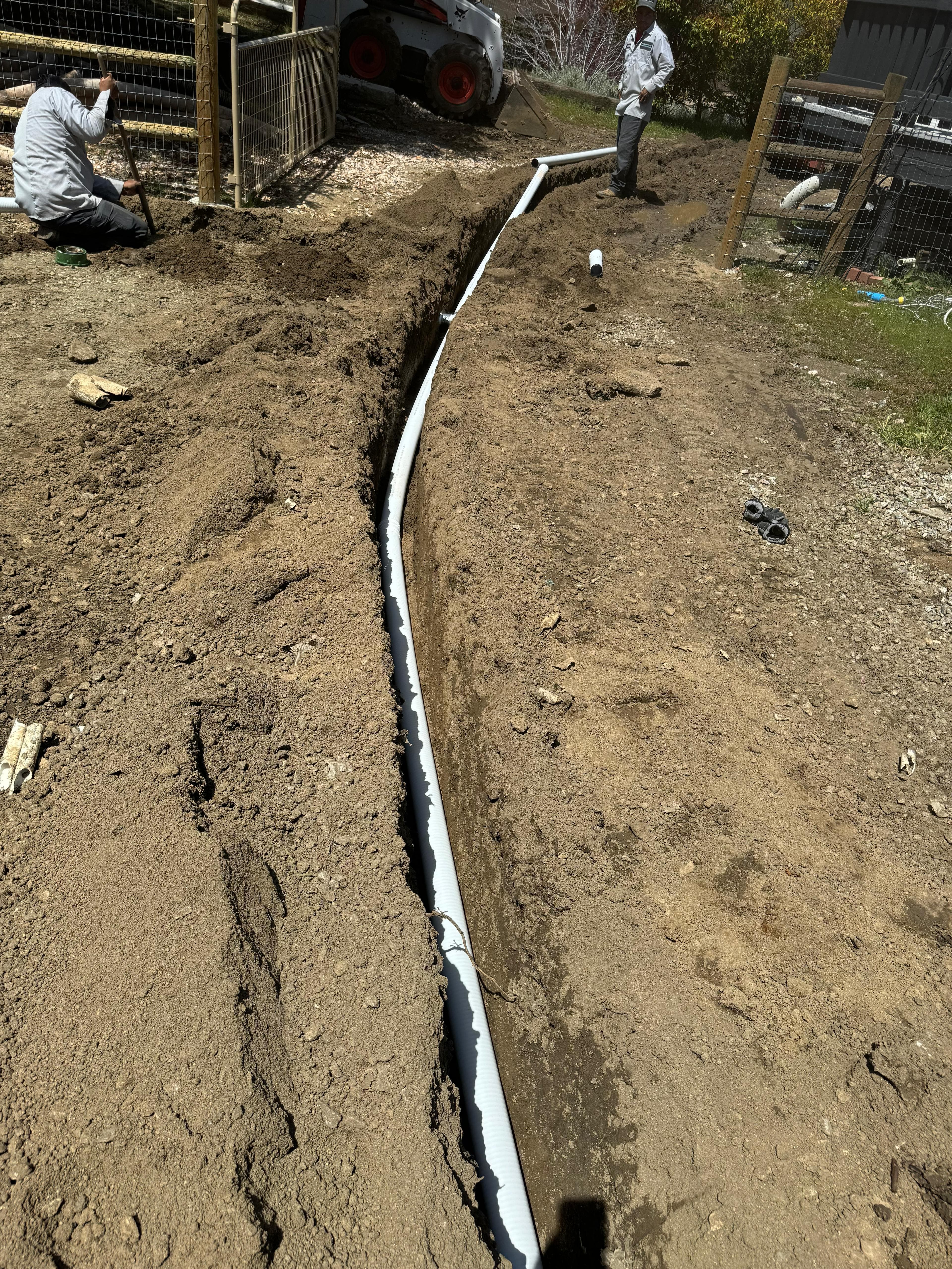 Long white pipe laid in a narrow dirt trench with workers and machinery nearby.