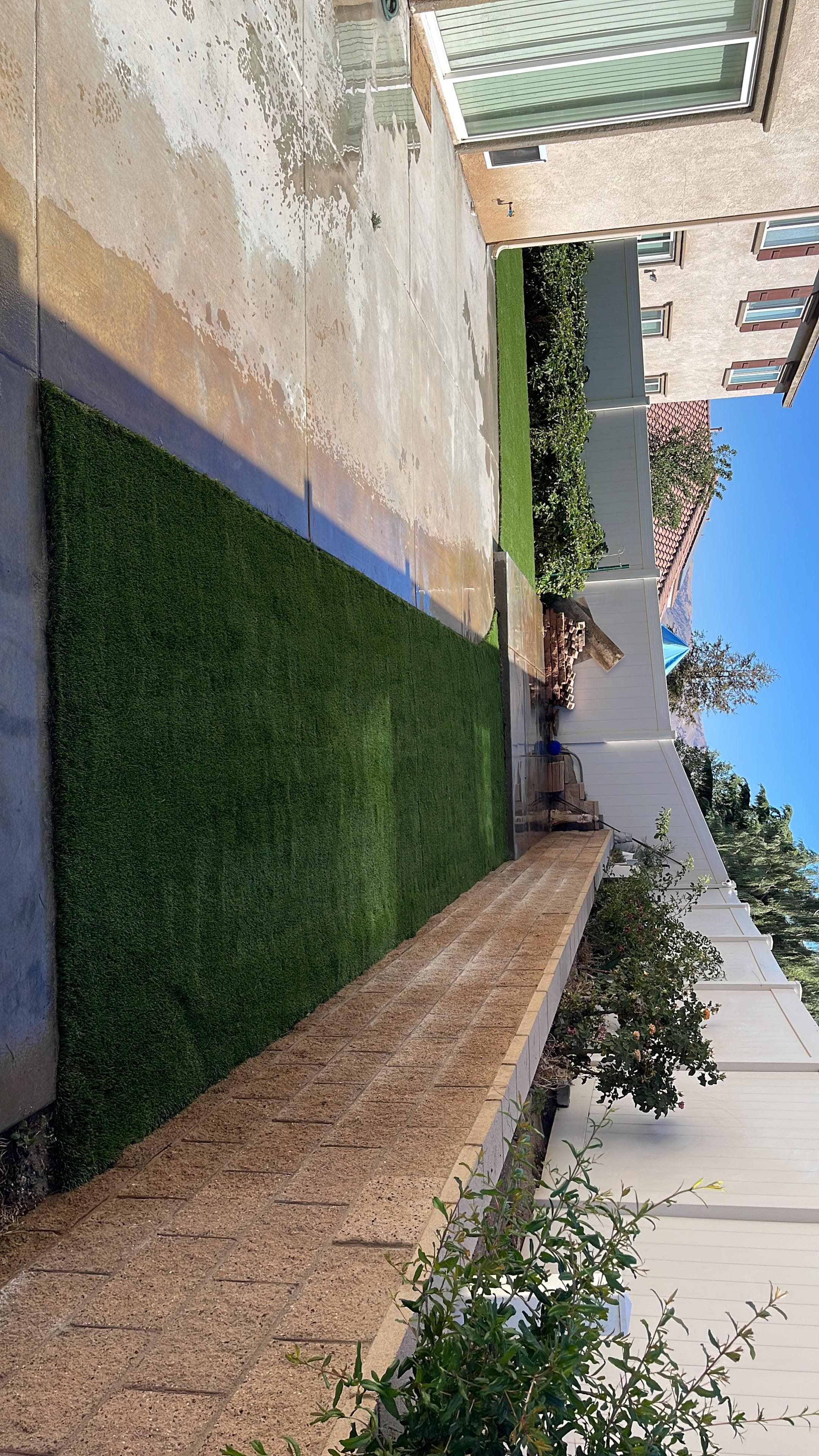 Green artificial turf strip alongside a wet concrete patio and a tan brick wall.