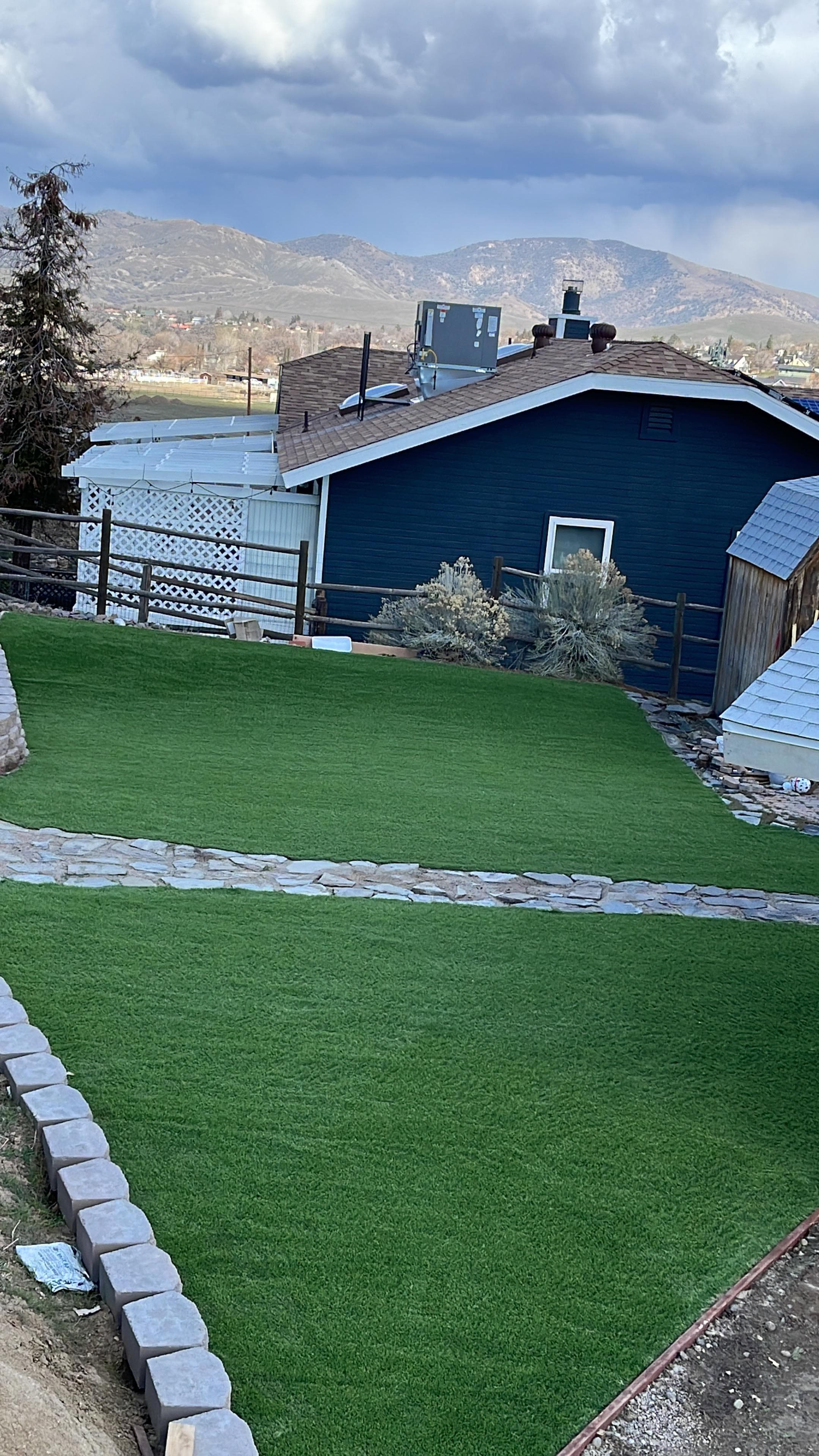 Lush green lawn with a stone path, a blue house, and mountains in the background.