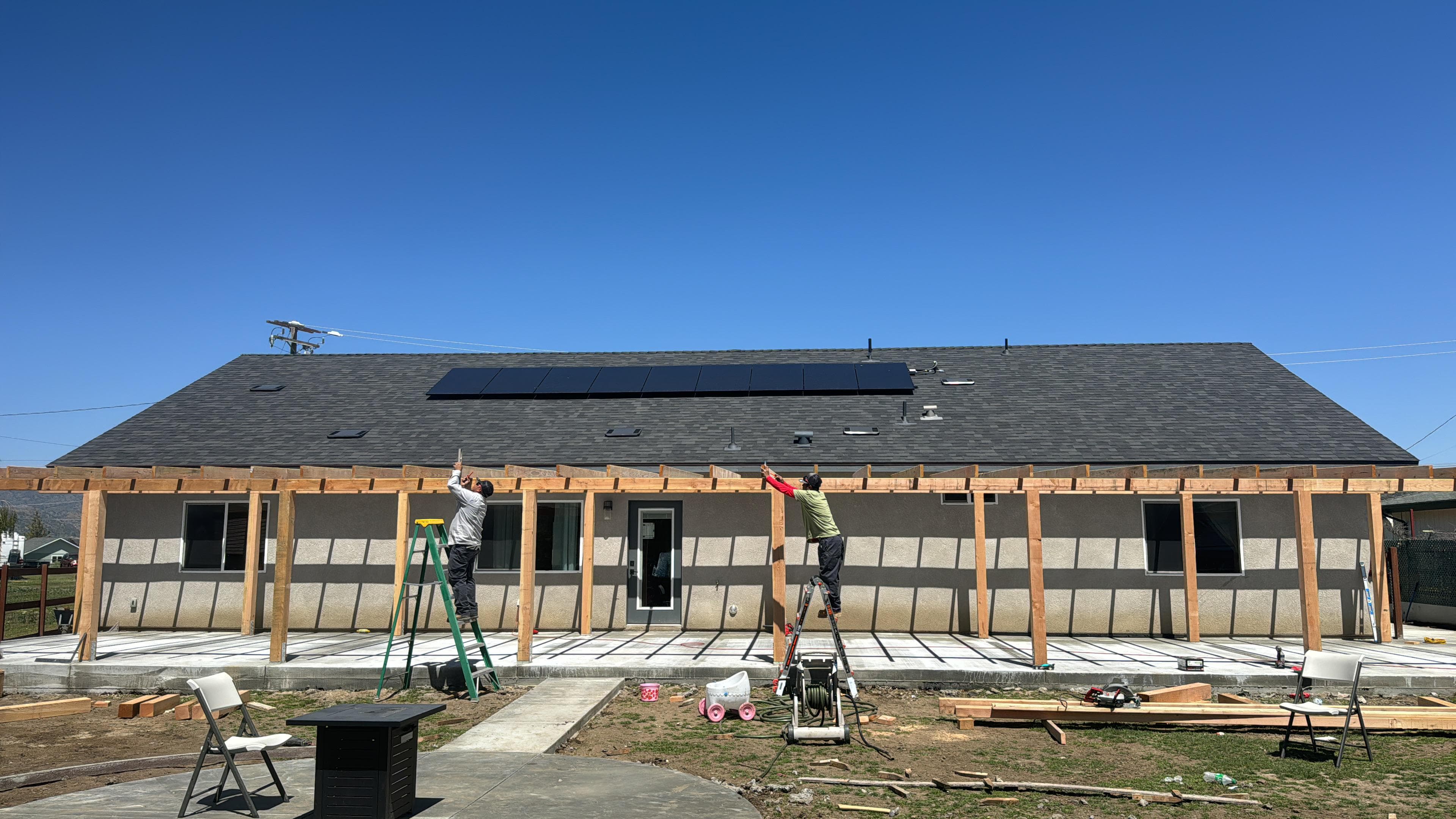 Two workers on ladders construct a wooden pergola frame against a house with solar panels.